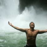 A joyful, shirtless man with his arms outstretched standing in front of a waterfall.
