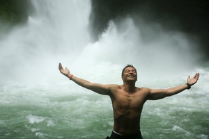 A joyful, shirtless man with his arms outstretched standing in front of a waterfall.