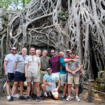 A group of tourists posing in front of an ancient temple overgrown with large tree roots.