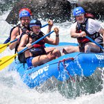 Four individuals wearing life vests and helmets white water rafting on a turbulent river.