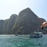 A person performing a backflip off a boat near a rocky island.