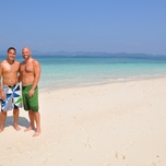 Two men standing side by side on a sunny beach with clear turquoise waters in the background.
