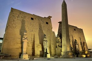 Stone wall with statues and a tall obelisk with luxor temple in the background.