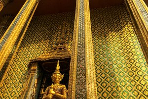 A golden statue of a guardian figure stands before the ornate facade of a temple adorned with intricate patterns and decorations, under a clear sky.