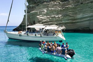 Show thumbnail preview A group of people on a dinghy next to a sailboat anchored near a cliff.
