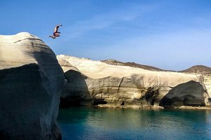 Person cliff diving into a serene blue sea near eroded rock formations.