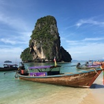 Traditional long-tail boats moored on a clear day at a beach with a limestone karst formation in the background.