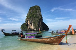 Traditional long-tail boats moored on a clear day at a beach with a limestone karst formation in the background.