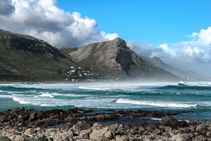 Rocky shoreline with rolling waves and a backdrop of rugged mountains under a cloudy sky.