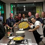 A group of smiling individuals posing with a large pan of paella, some wearing aprons indicating a cooking class setting.