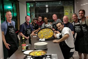 A group of smiling individuals posing with a large pan of paella, some wearing aprons indicating a cooking class setting.