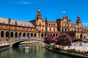 A sunny view of the plaza de españa in seville with a bridge over the canal, blossoming trees, and visitors exploring the grounds.