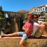 Two people posing in front of an ancient bridge with a backdrop of historic buildings on a sunny day.
