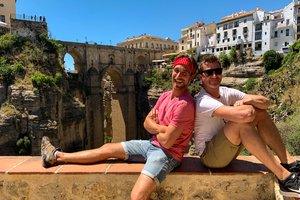 Two people posing in front of an ancient bridge with a backdrop of historic buildings on a sunny day.