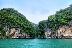 Limestone cliffs bordering a tranquil turquoise sea inlet under a cloudy sky.