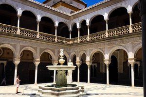 A courtyard with an ornate fountain, surrounded by arches and intricate stonework, with a person standing to the side.