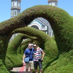 Two men smiling and standing arm-in-arm under a heart-shaped archway with a church in the background.
