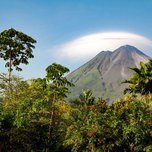 A volcano looms behind lush greenery under a clear sky with a lenticular cloud formation